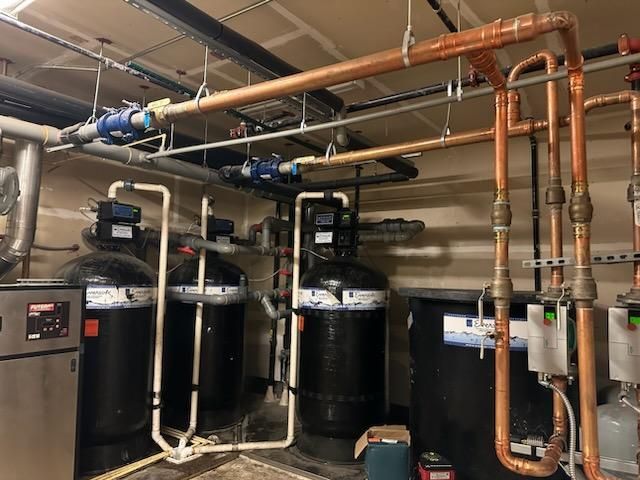 Plumbing equipment installed in a basement, including tanks, pipes, and valves. Copper pipes and black tanks are visible.