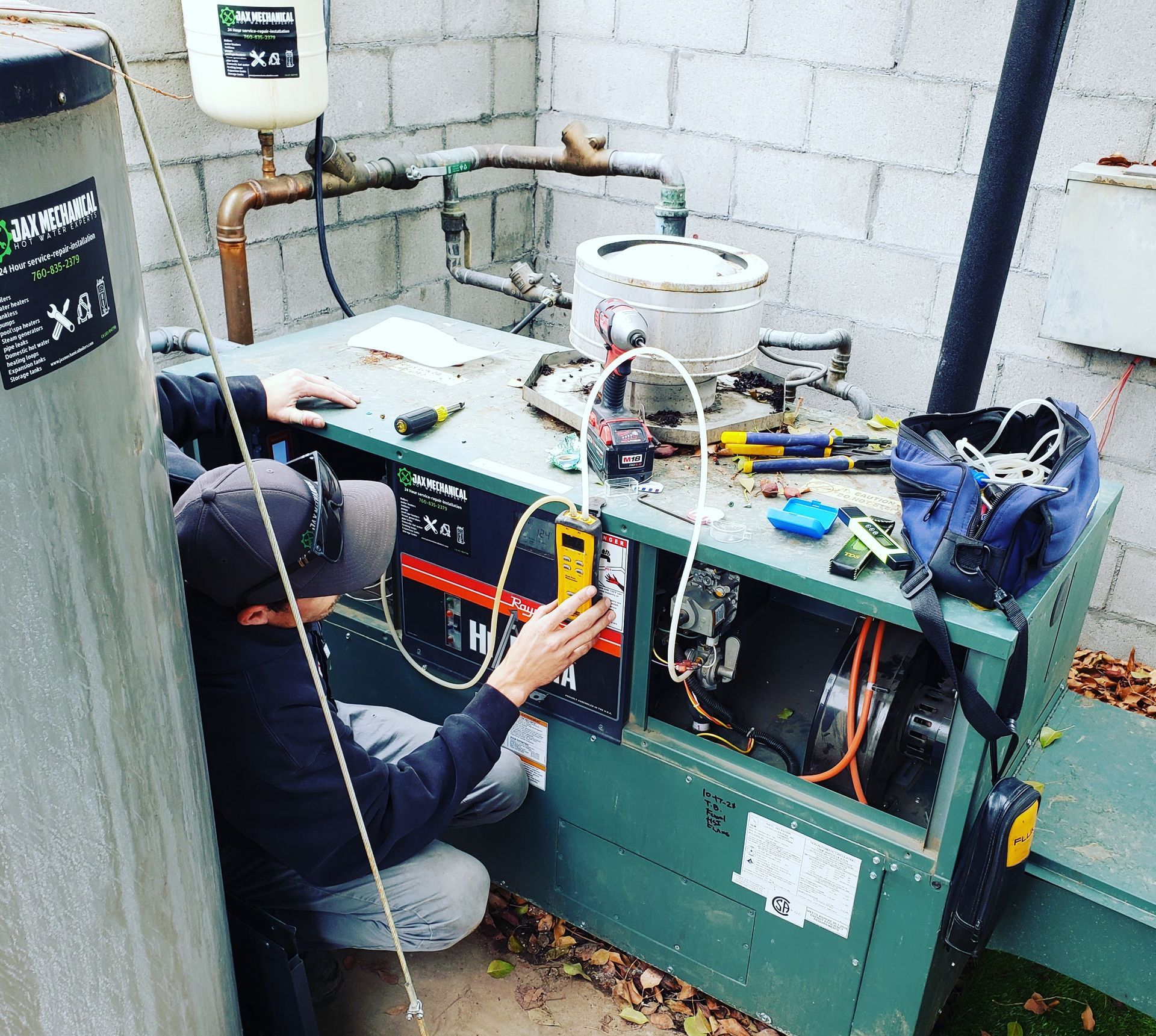 A person in a cap kneels, using a meter on a heating unit, outdoors next to a water heater.