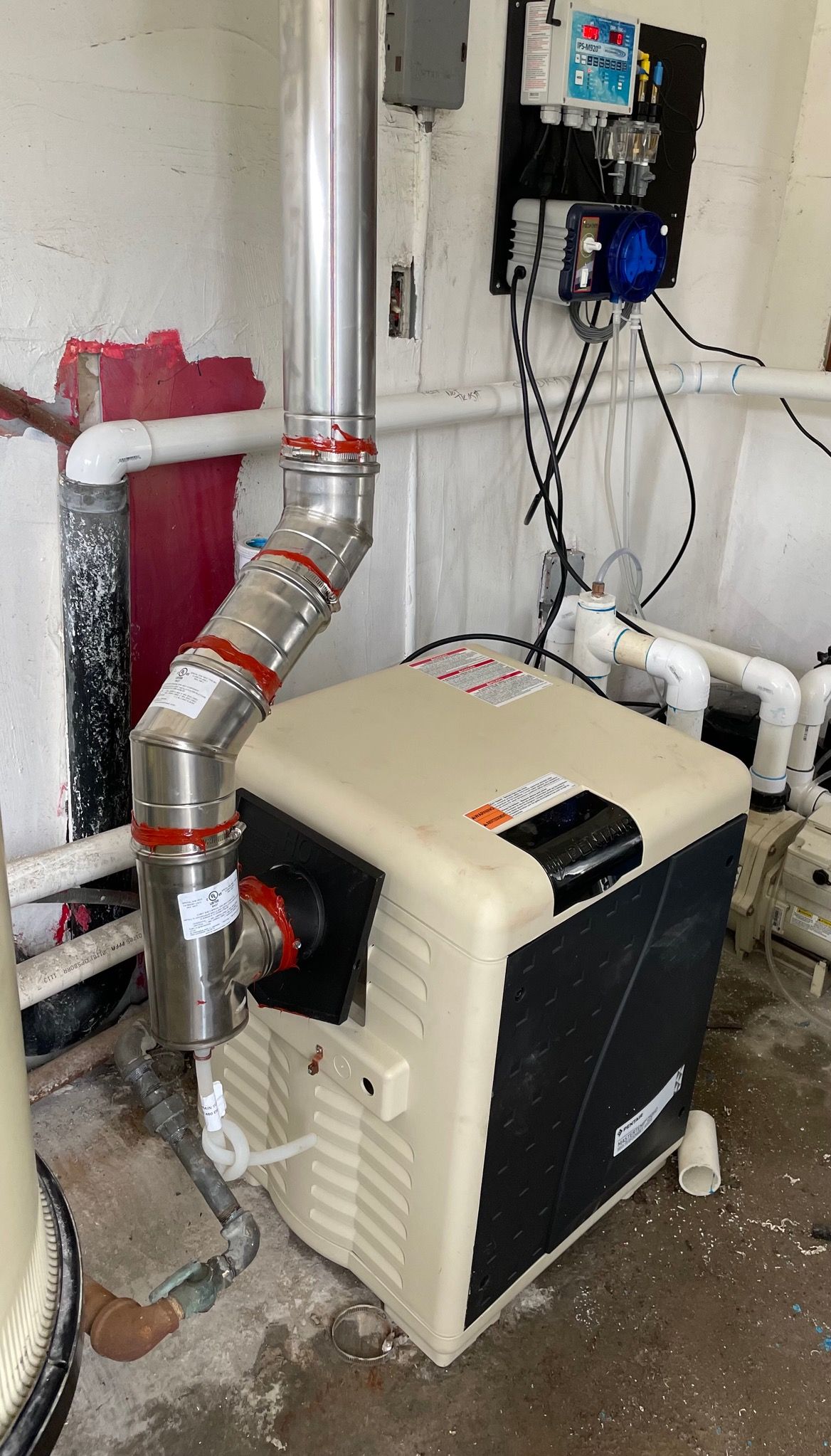 Pool heater with stainless steel exhaust pipe against a white wall with associated electrical equipment.