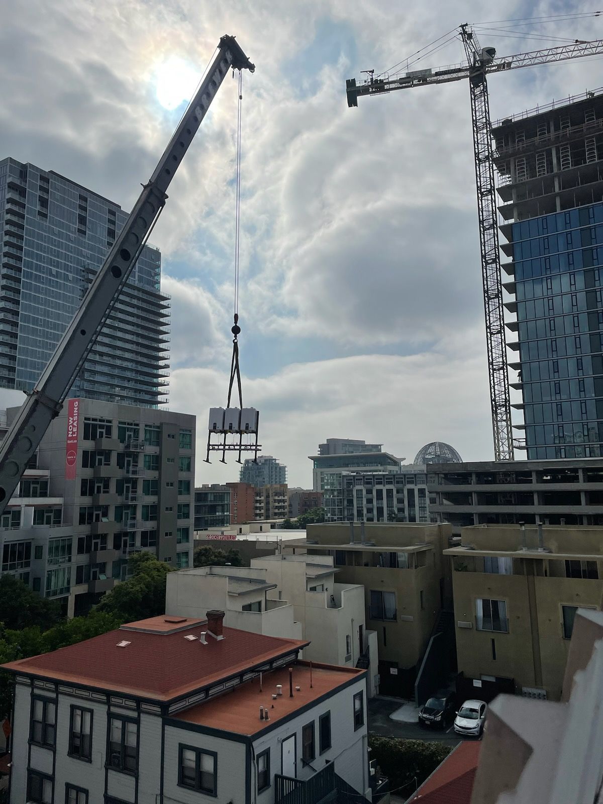 A large crane lifts water heaters over city buildings, with a taller construction crane in the background under a cloudy sky.