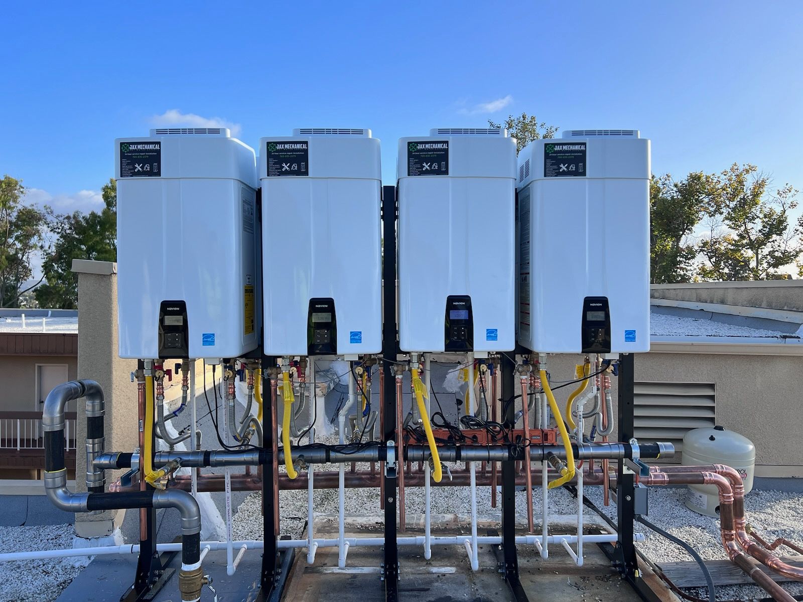 Four white tankless water heaters installed on a rooftop, connected by pipes and electrical wiring.