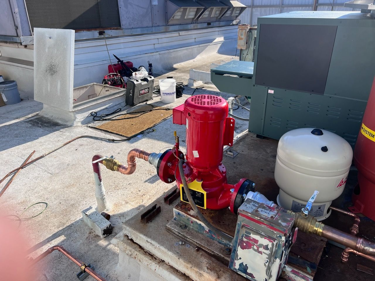 A red pump and associated plumbing on a rooftop. Other equipment, tools, and a water tank are also visible.