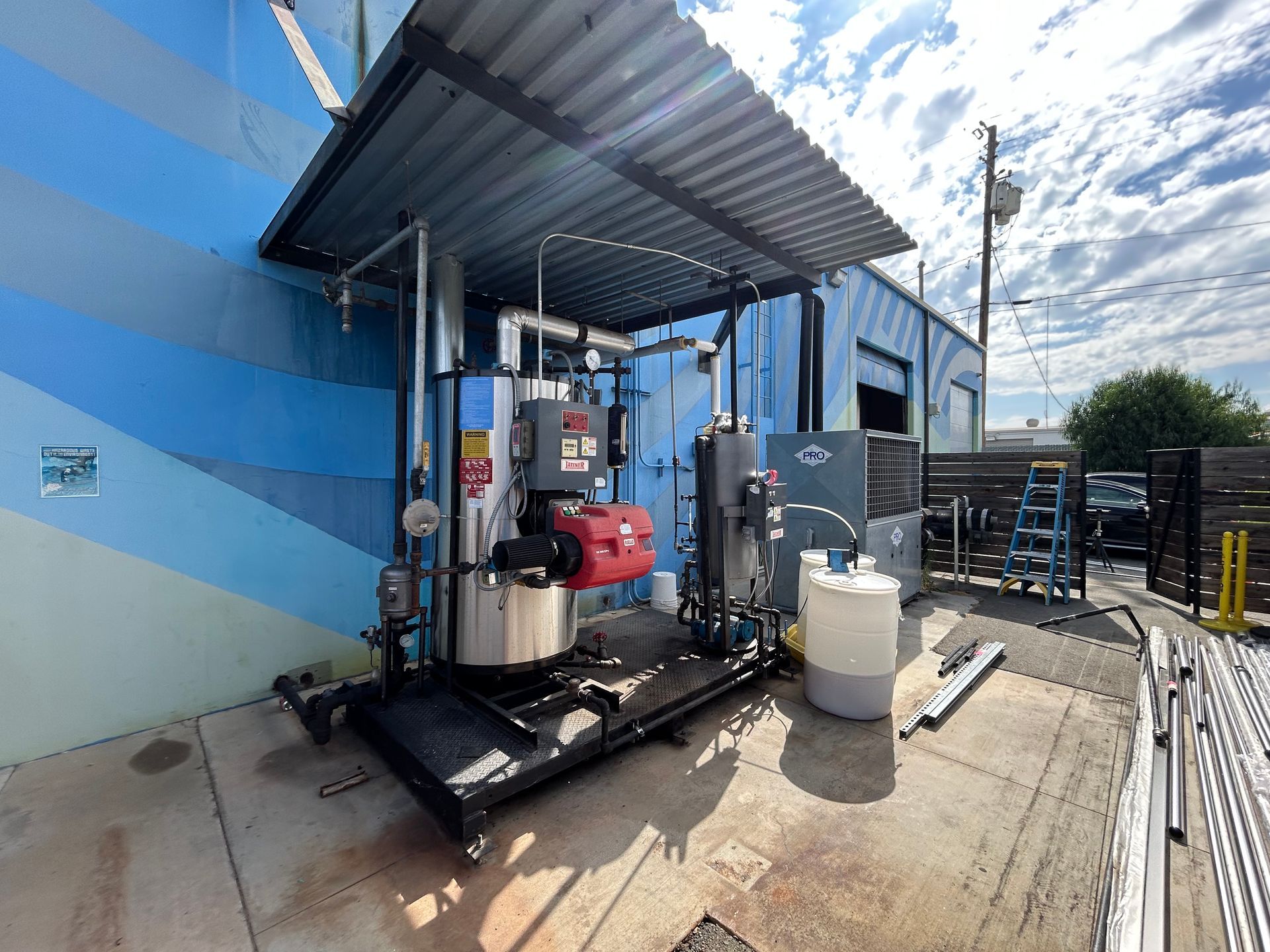 A large industrial boiler system outdoors with a corrugated metal awning, mounted against a blue mural-covered wall.