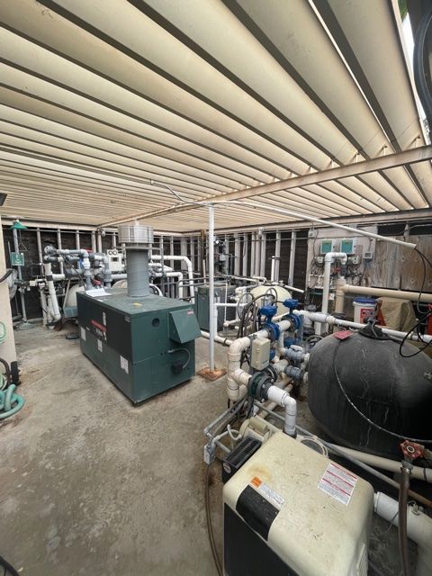 A cluttered room with pool equipment, including a large heater, pipes, and tanks under a slatted ceiling.