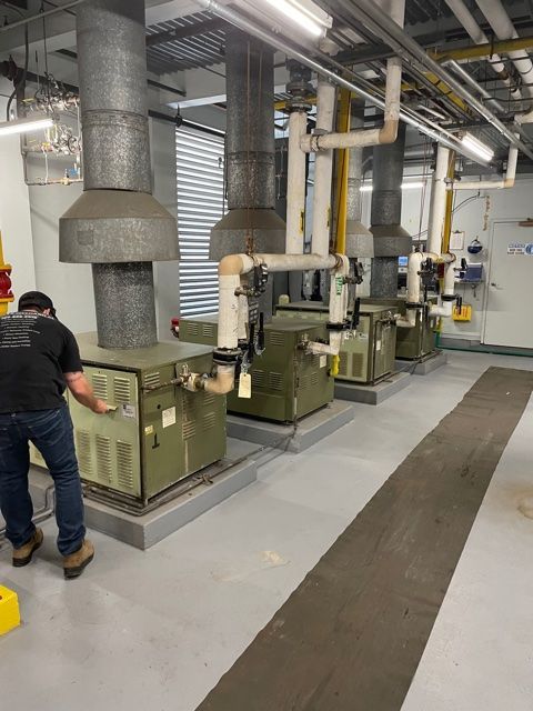 A person in a black shirt inspects machinery in an industrial setting with several boilers and vents.