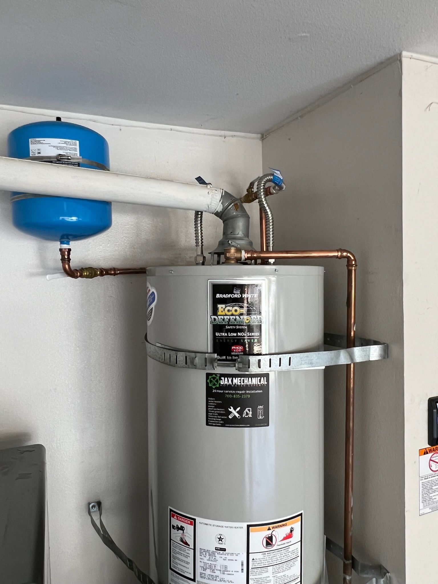 A tall, gray water heater with copper pipes and an expansion tank attached in a corner of a room.