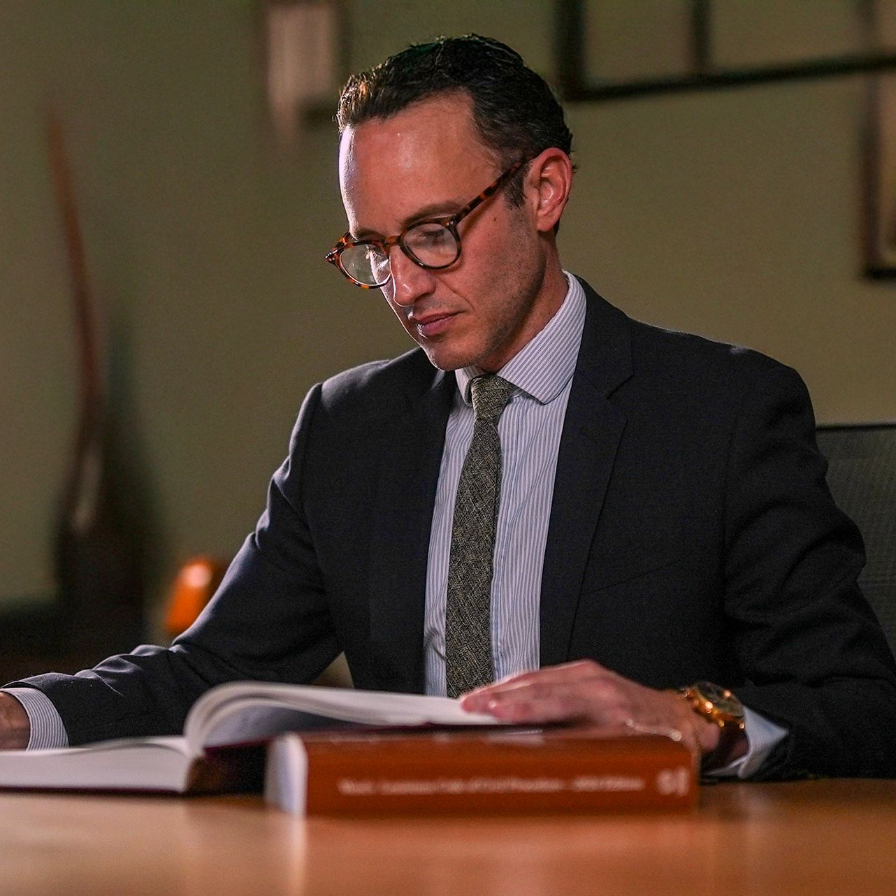 Man in a suit, reading a book, holding glasses, at a table, likely in an office.