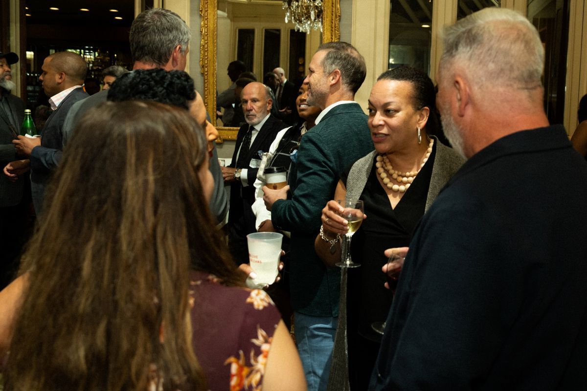 People at an event, some holding drinks, in a well-lit room. Woman in a dark dress speaks with a man.