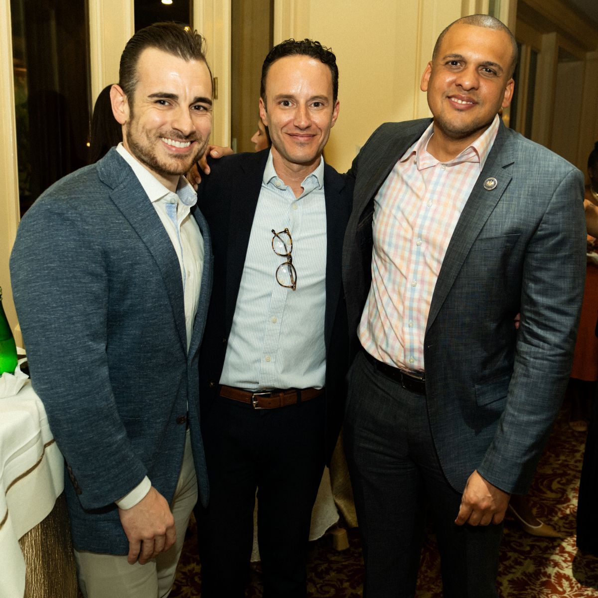 Three men smiling at a gathering, wearing suits, in a warmly lit room.