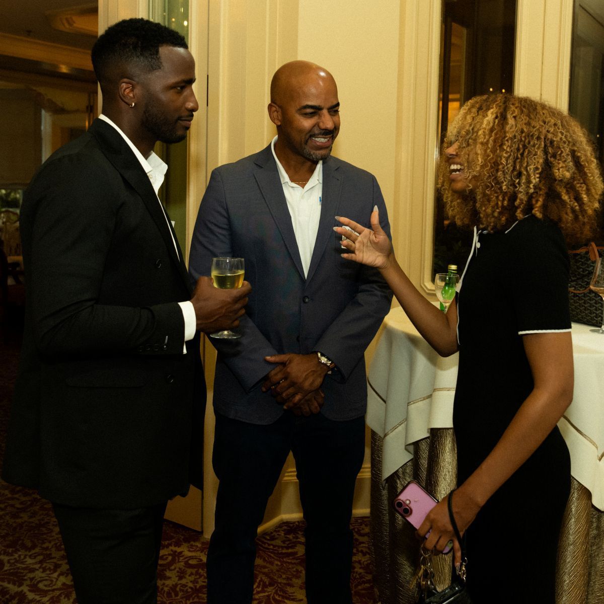 Three people in a room, two men in suits and a woman in a black dress, chatting and smiling.