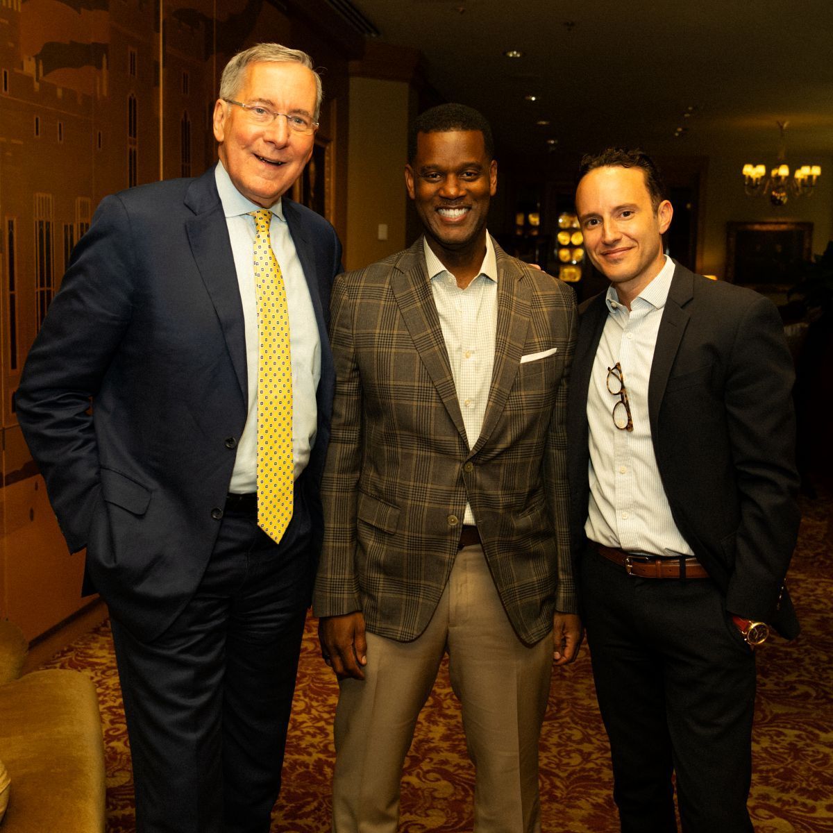 Three men smiling; one in a blue suit, another in a checkered blazer, and the last in a black suit, indoors.