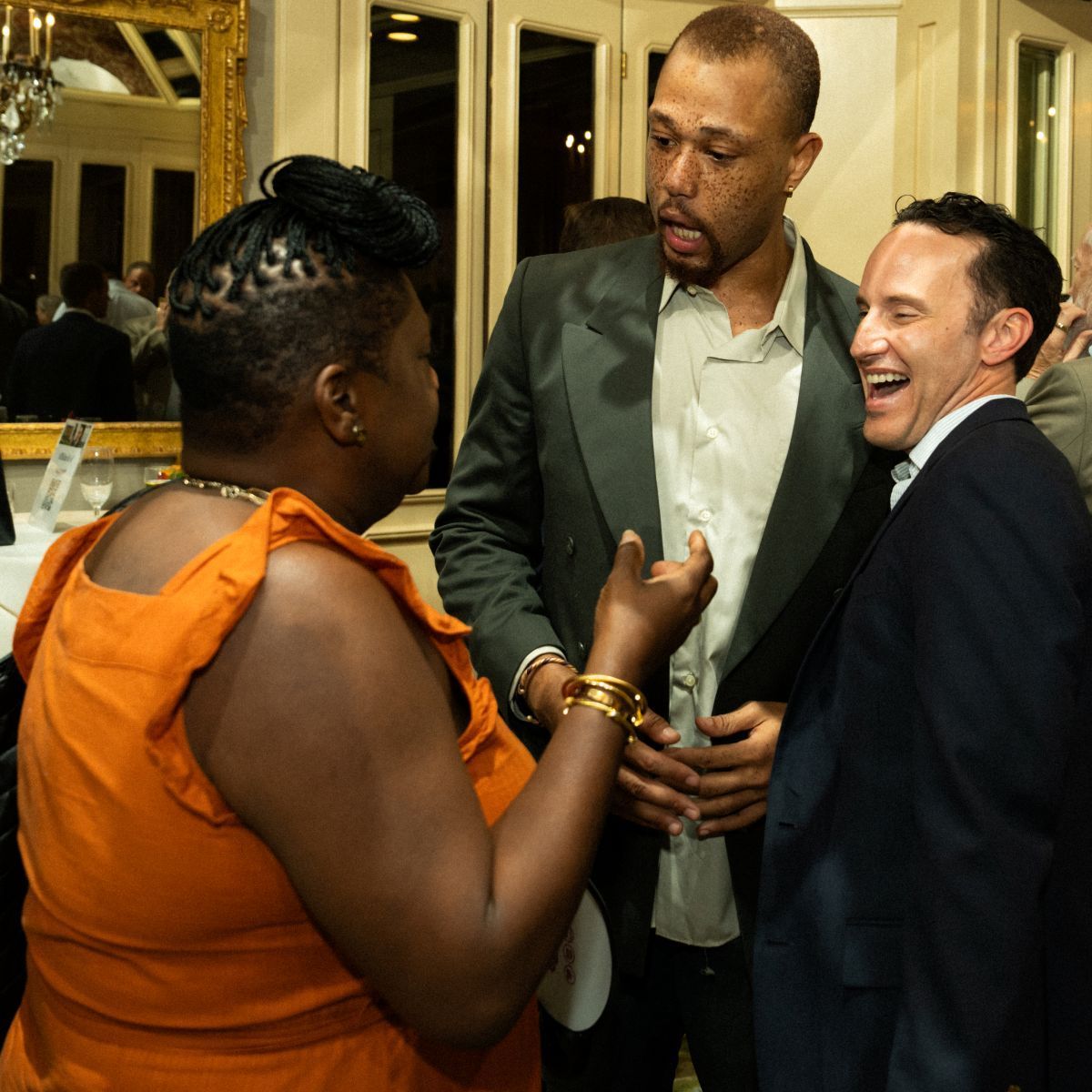 Three people conversing; a woman in orange gestures; two men listen, one laughs, all in a room with a gold-framed mirror.