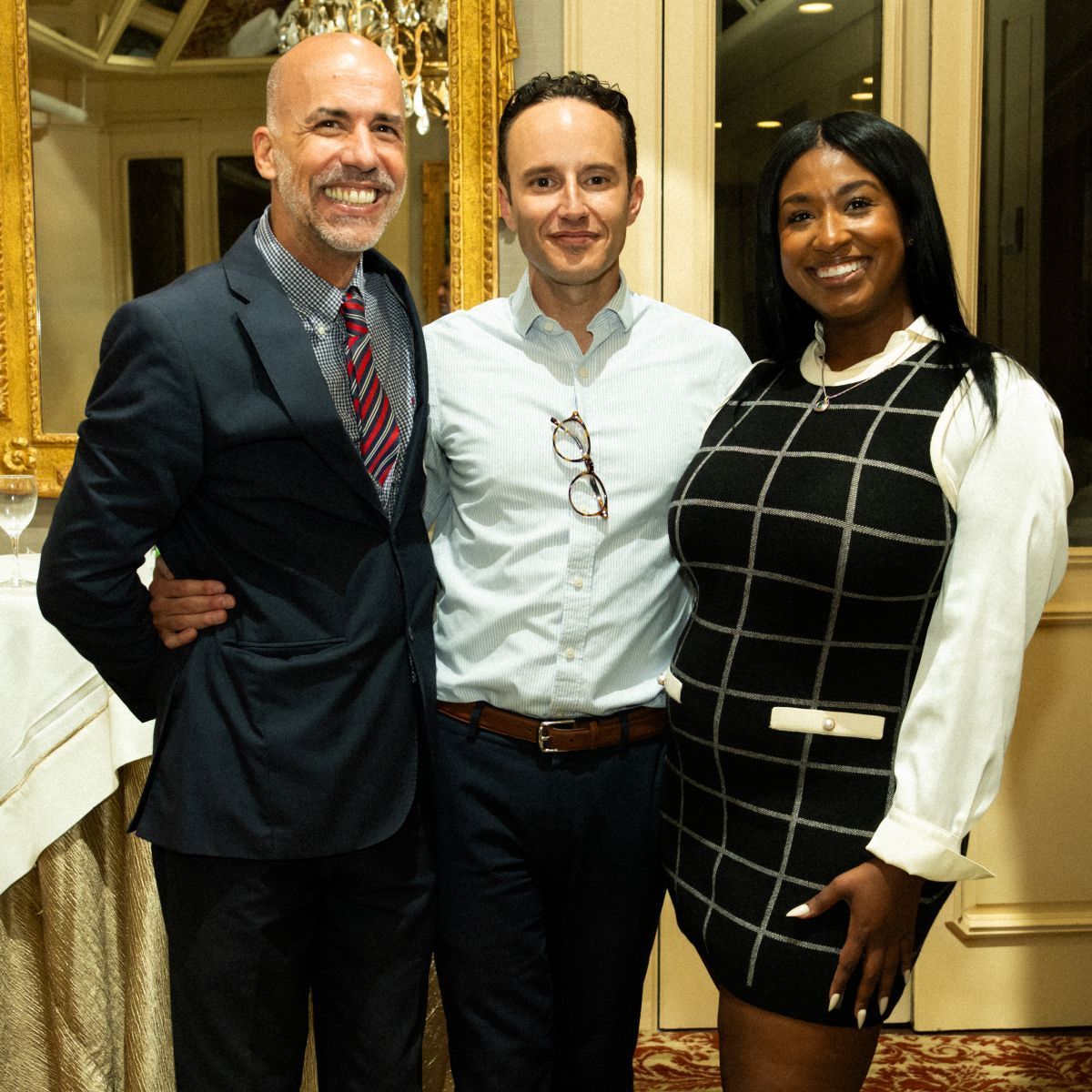 Three people smiling, posing for a photo. A man in a suit, a man with glasses, and a woman in a plaid dress.