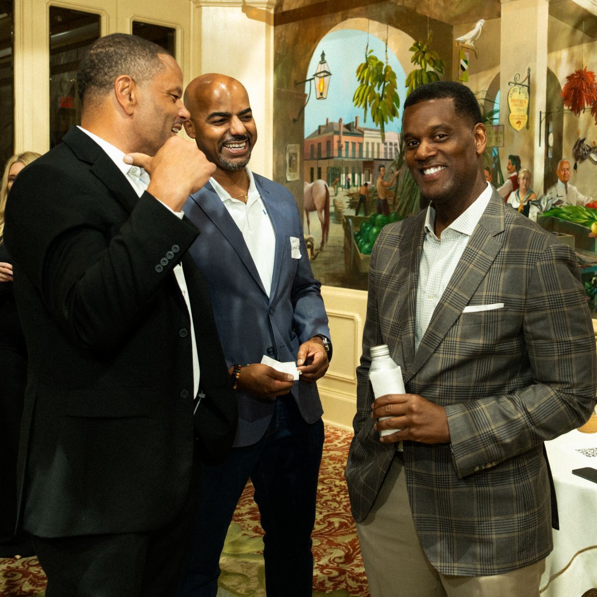 Three men in suits conversing in a warmly lit room with a painted mural.