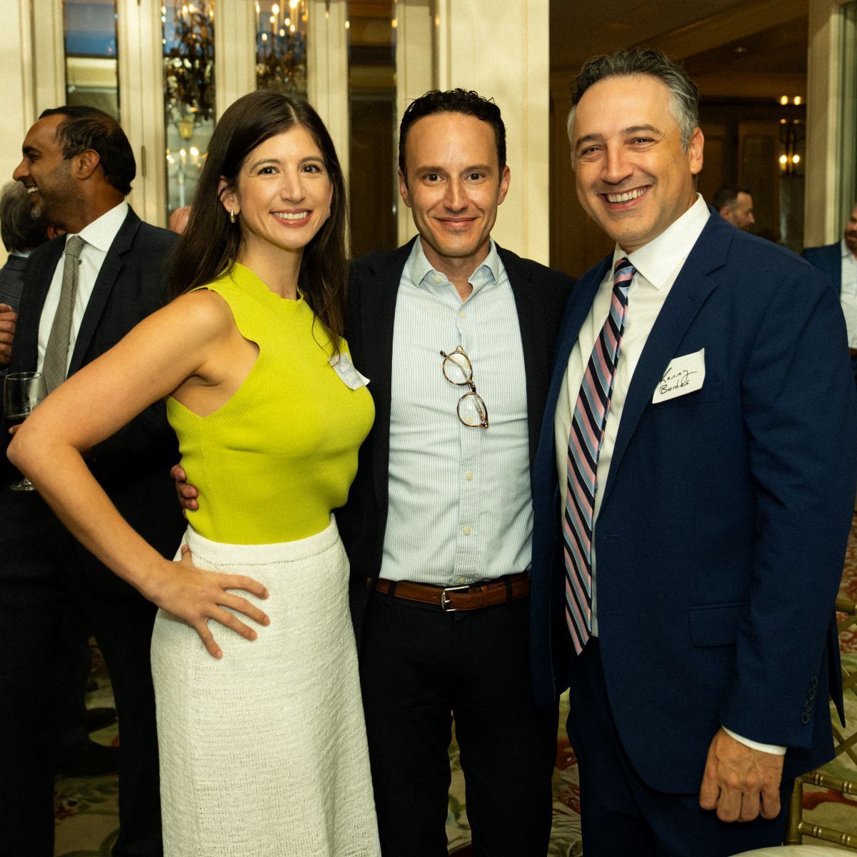 Three smiling people at event: woman in yellow top and white skirt, two men in suits.