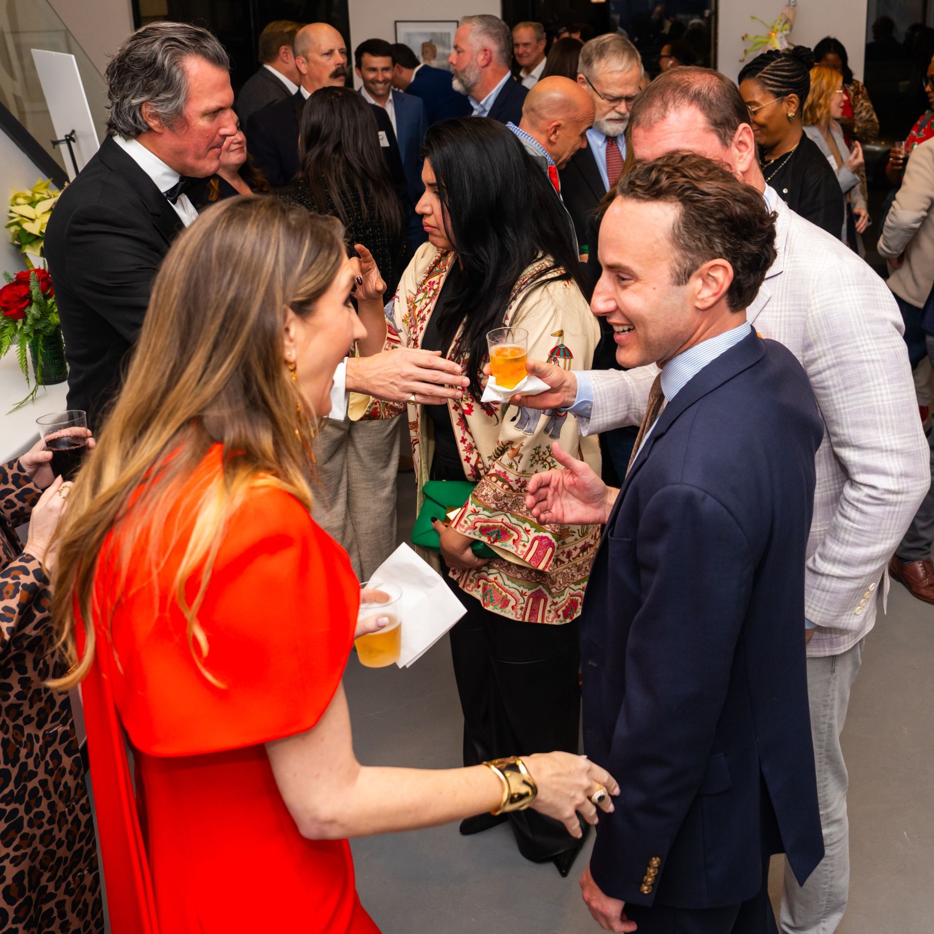 People socializing at an event, many in business attire. Woman in red dress, man in blue suit, interacting.