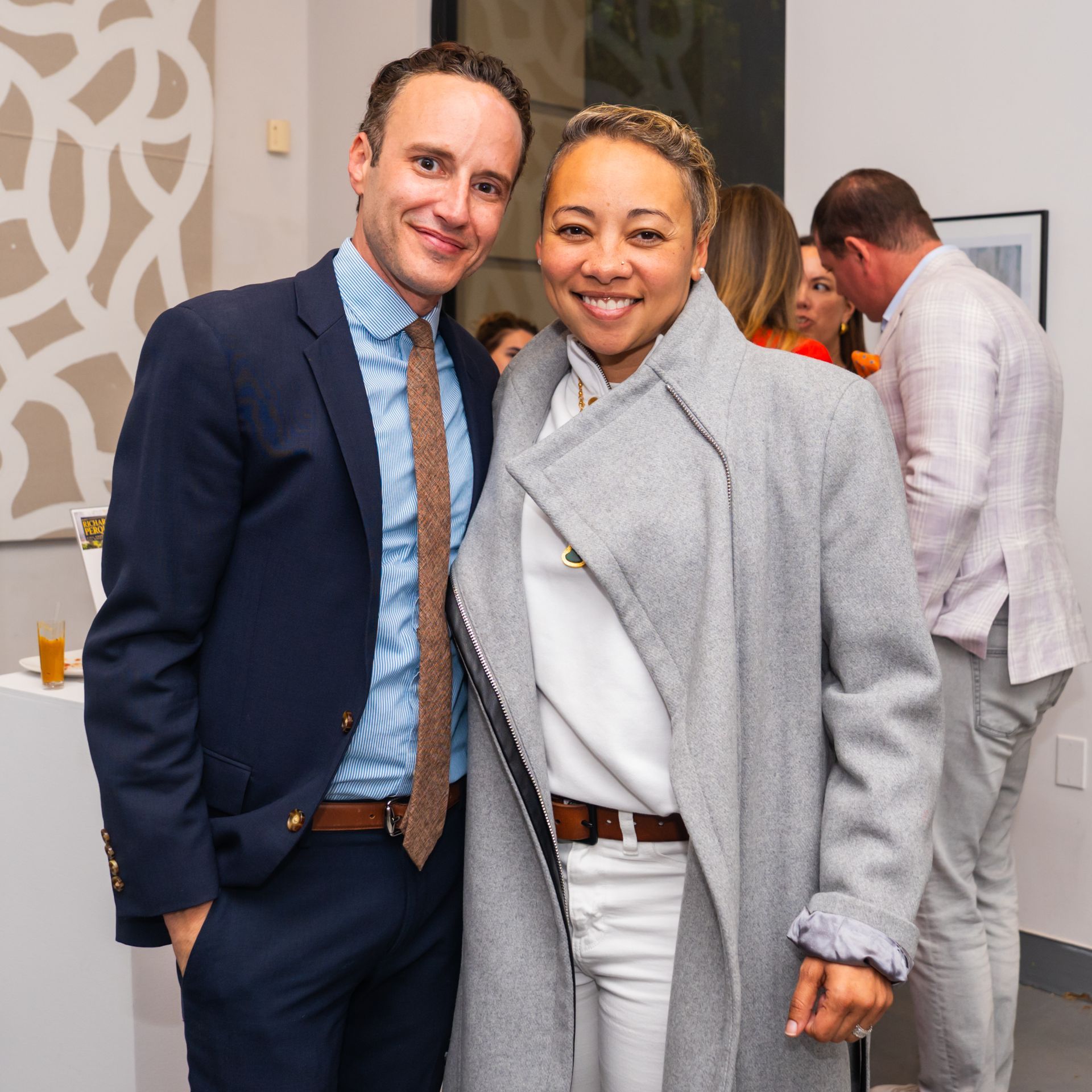 A man in a suit and a woman in a grey coat pose for a photo at an event.