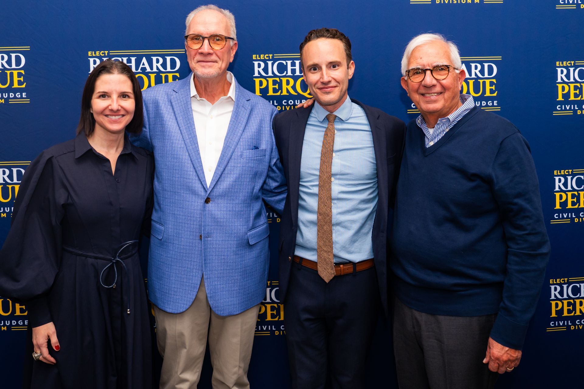 Four people posing in front of a navy backdrop with gold text. Woman in navy dress, three men in suits, smiling.
