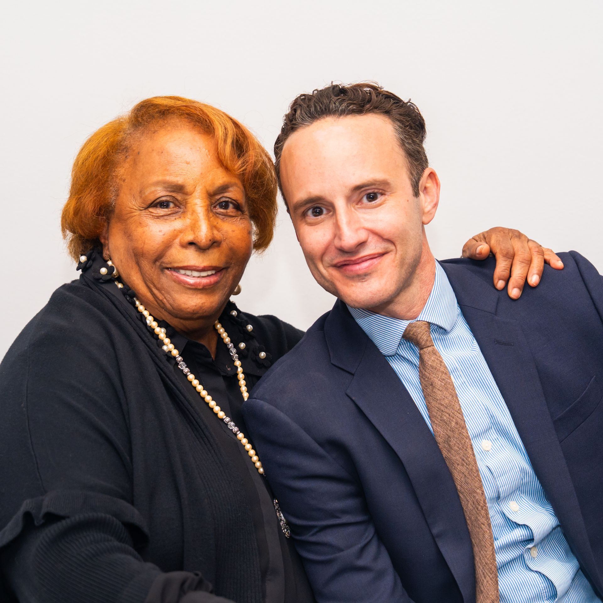 Woman and man smiling, close together, posing. Man has arm around woman. They are indoors.