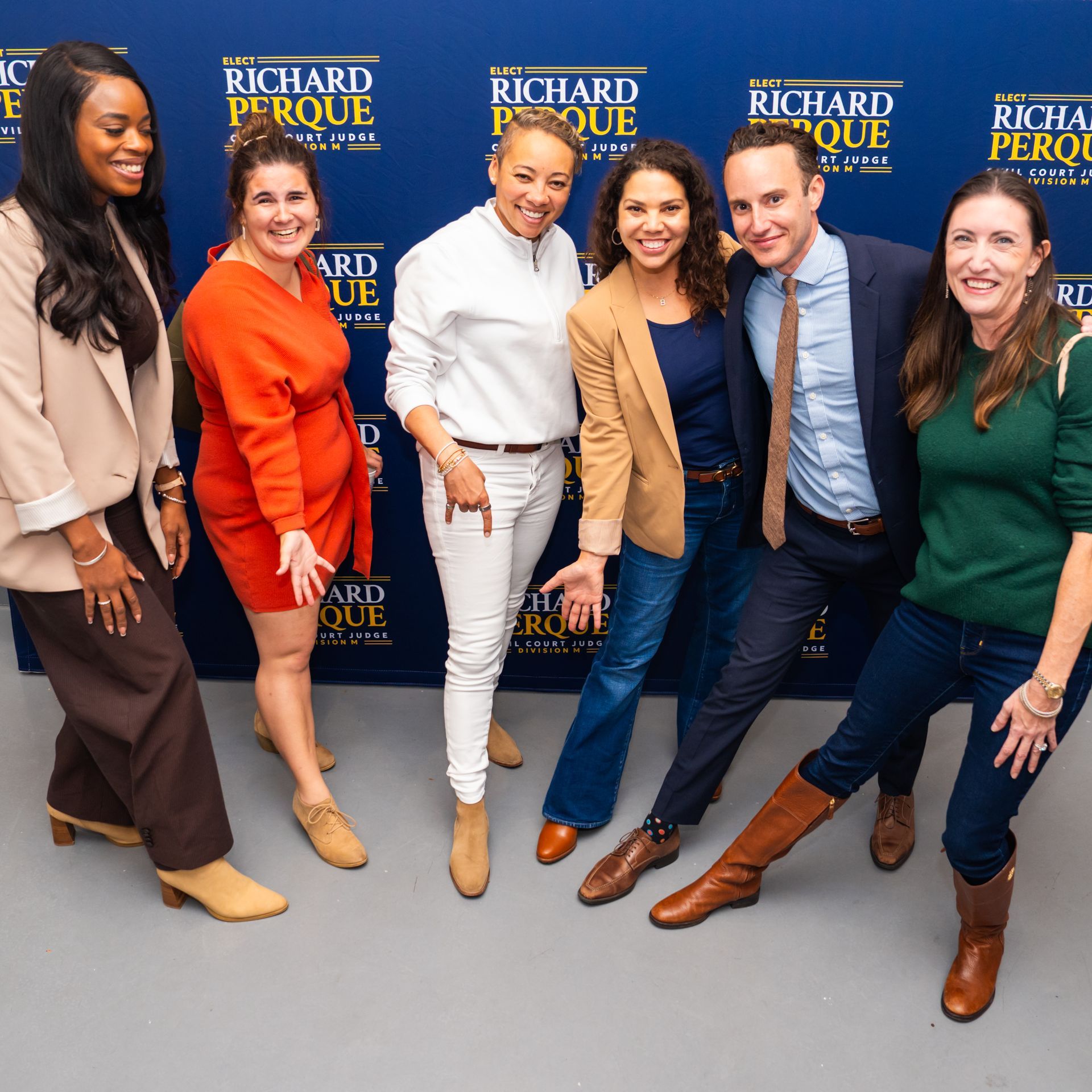 A group of seven people smiling and posing in front of a blue backdrop with text. They are wearing various outfits.
