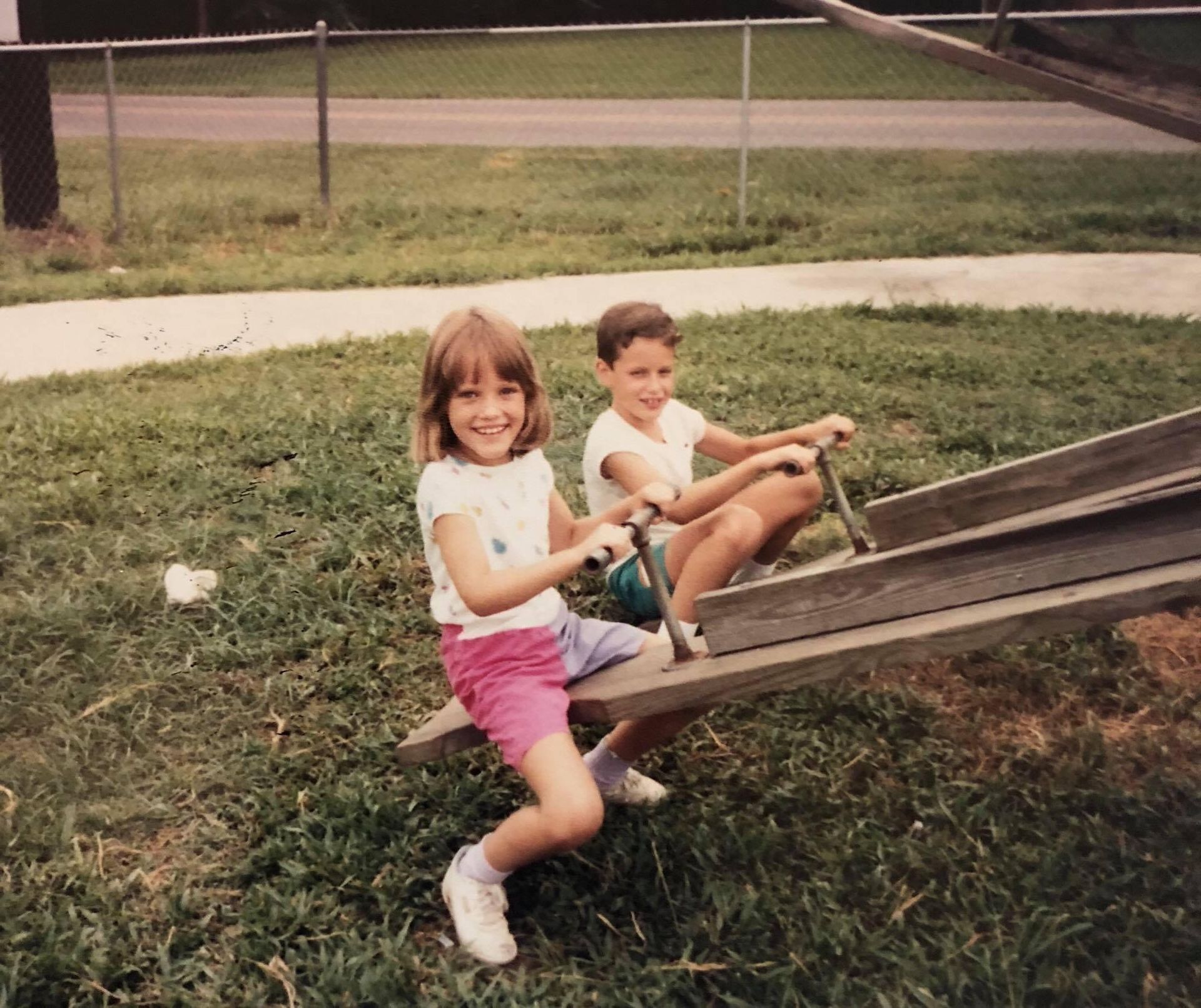 Richard and his sister as children on a wooden seesaw in a grassy area, smiling.