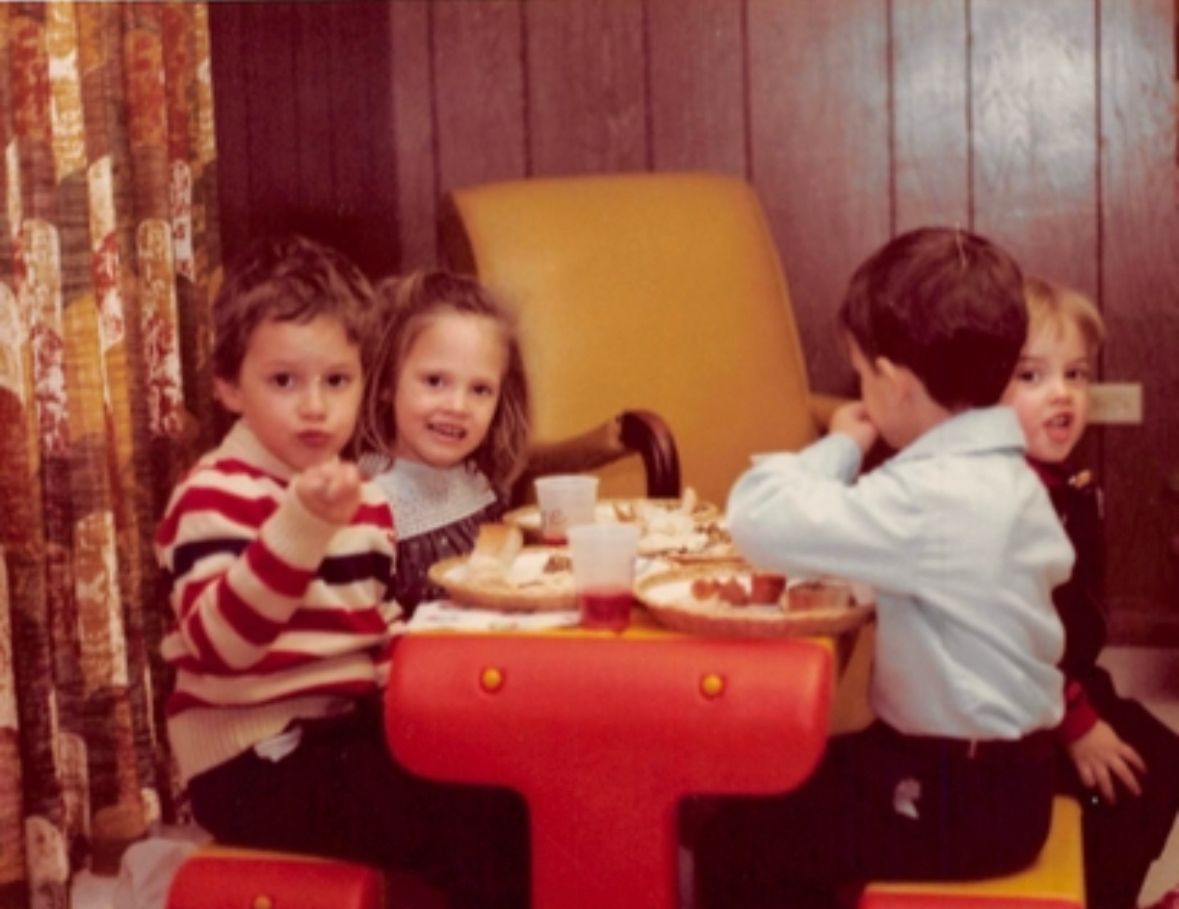 Four children at a red and yellow table, eating.  A striped shirt, a white blouse, and a blue shirt are visible. 