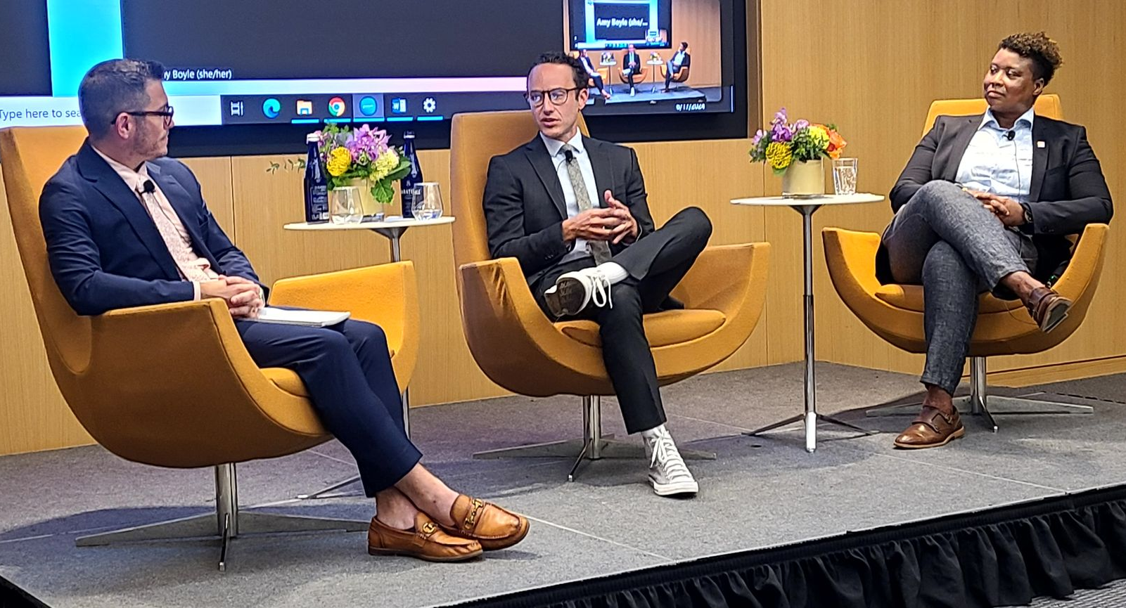 Richard Perque and two other people in modern chairs on a stage, engaged in discussion. The backdrop includes a screen and floral arrangements.
