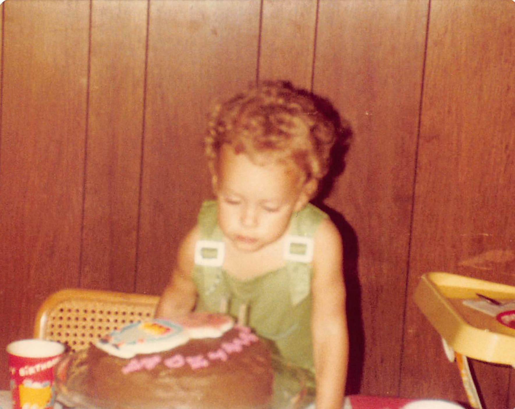 Child at a birthday cake, wearing a green dress. The child appears to be blowing on the cake.