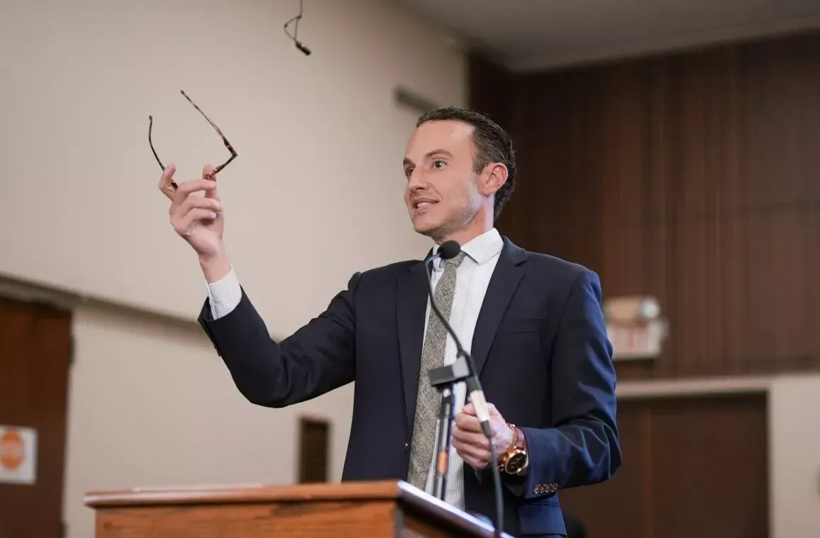 Man in suit holding glasses up in the air while speaking at a podium in a room.