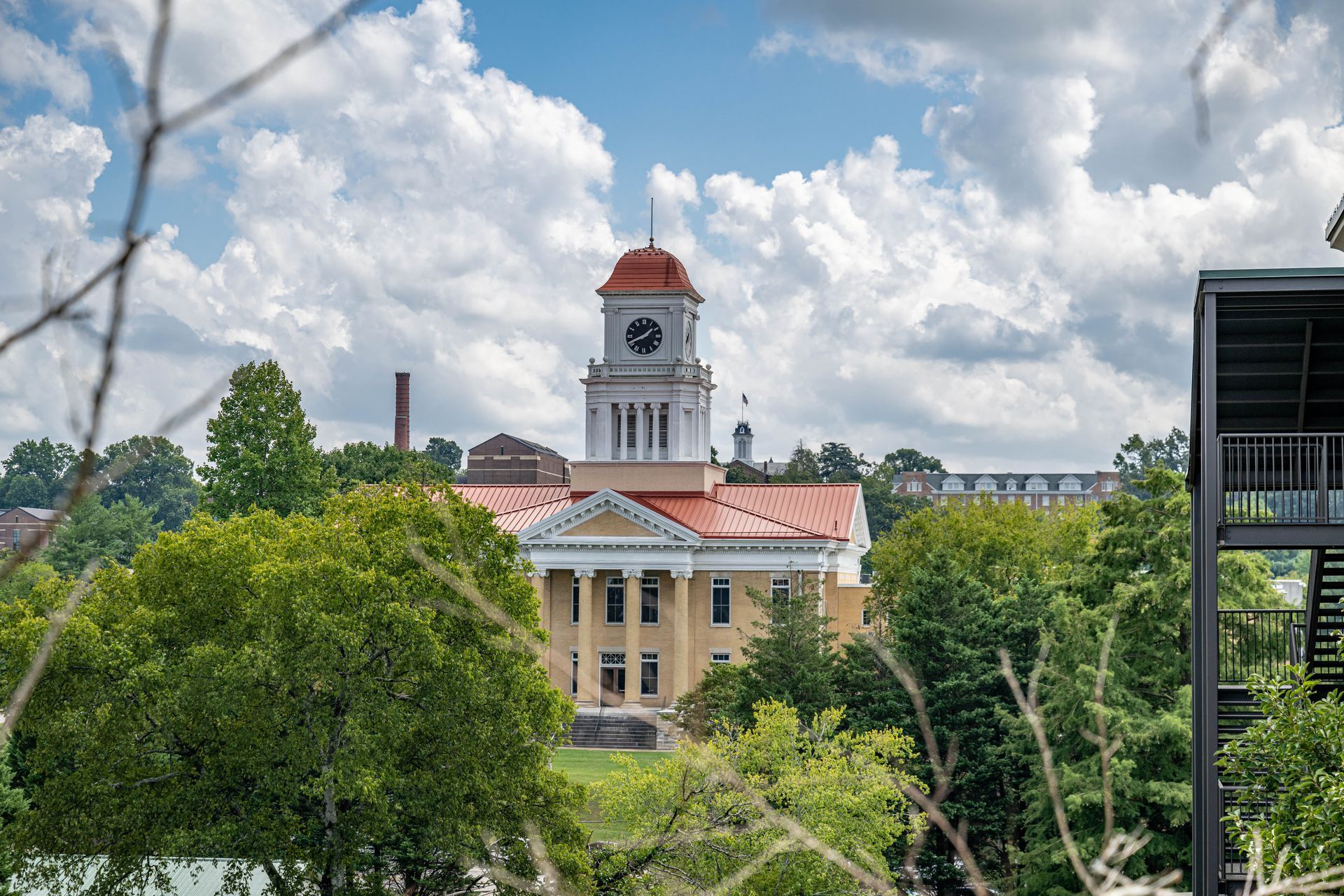 Clock tower of a classical building with columns, orange roof, and cloudy sky backdrop.