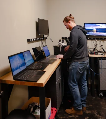 Person standing at a workstation with multiple monitors in a small office or studio.