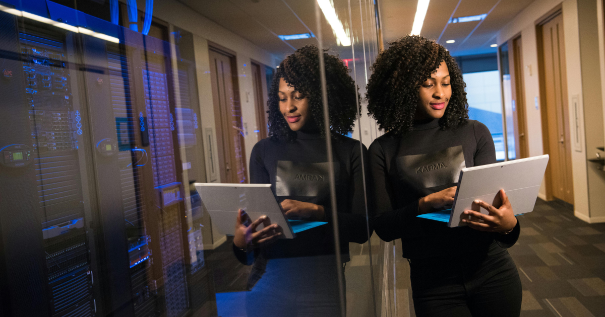 Woman with curly hair in black sweater, leaning against a glass wall while using a laptop in an office hallway.