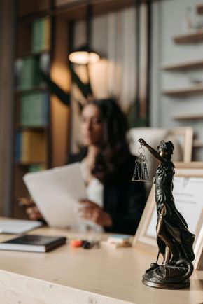 Statue of Lady Justice on desk in lawyer's office; woman reviewing documents in background.