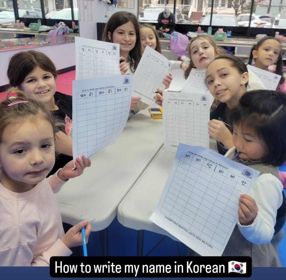 A group of young girls are sitting at a table holding papers that say how to write my name in korean