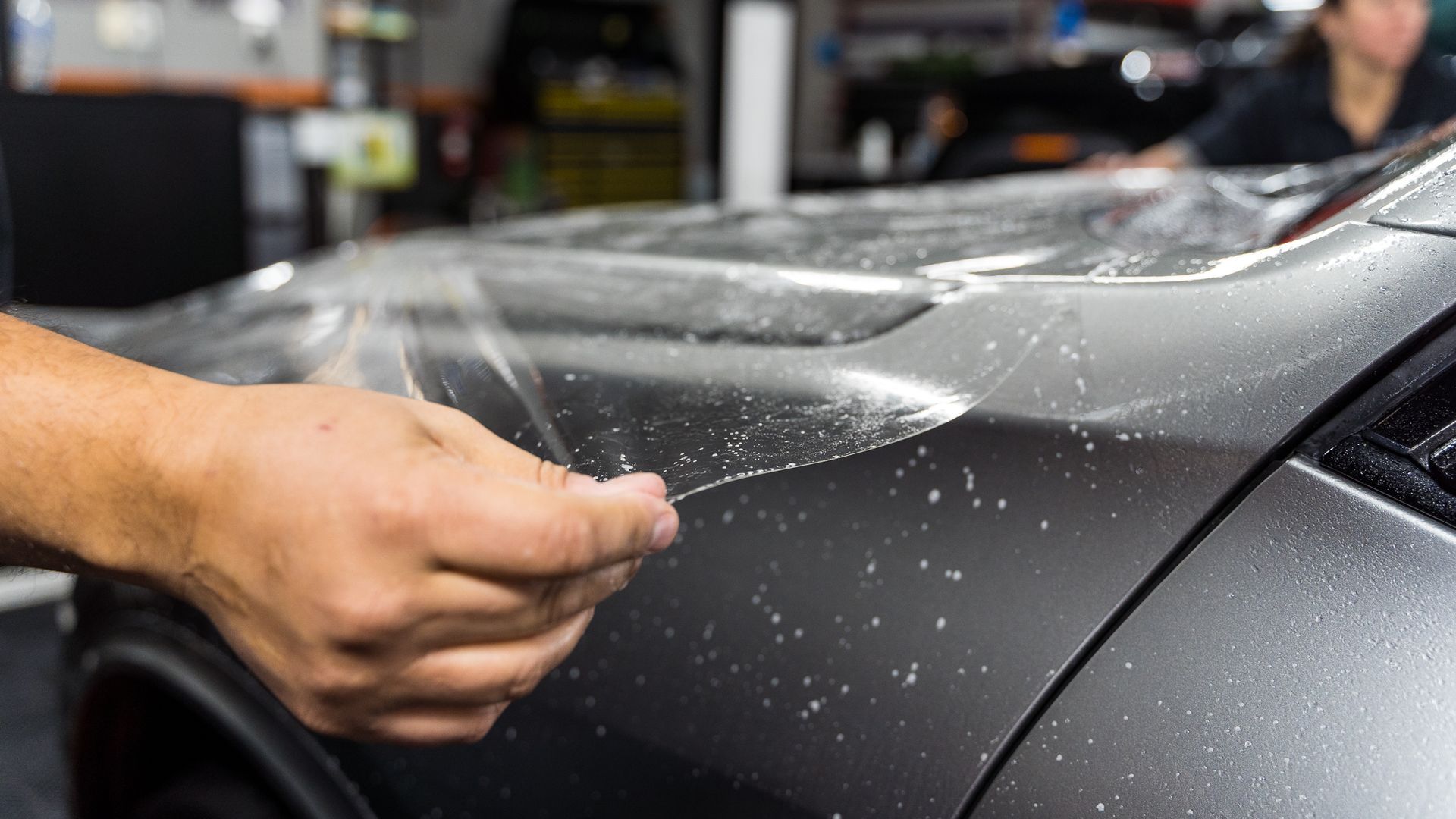 Person applying a clear protective film to the gray hood of a car in a workshop.