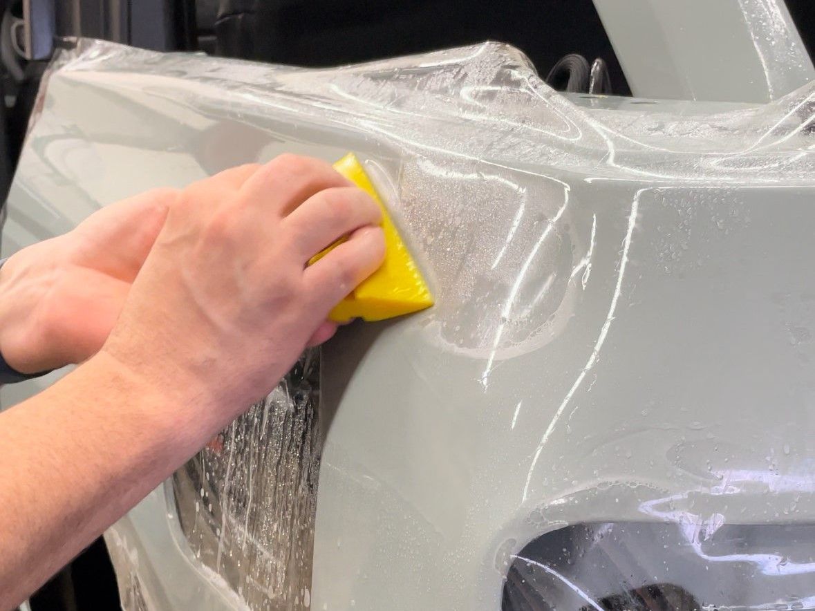 Person using a yellow squeegee to apply clear protective film to the front bumper of a light gray car.