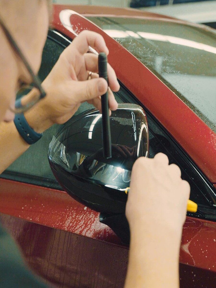 Person applying film to a car's black side mirror with a squeegee and tool; red car visible.