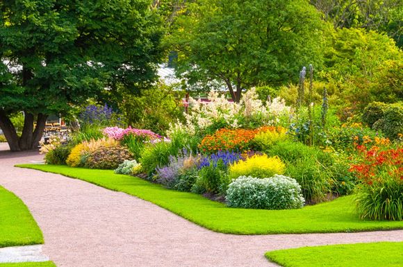 Curving gravel path borders vibrant flowerbeds in a lush garden, with green grass and trees.