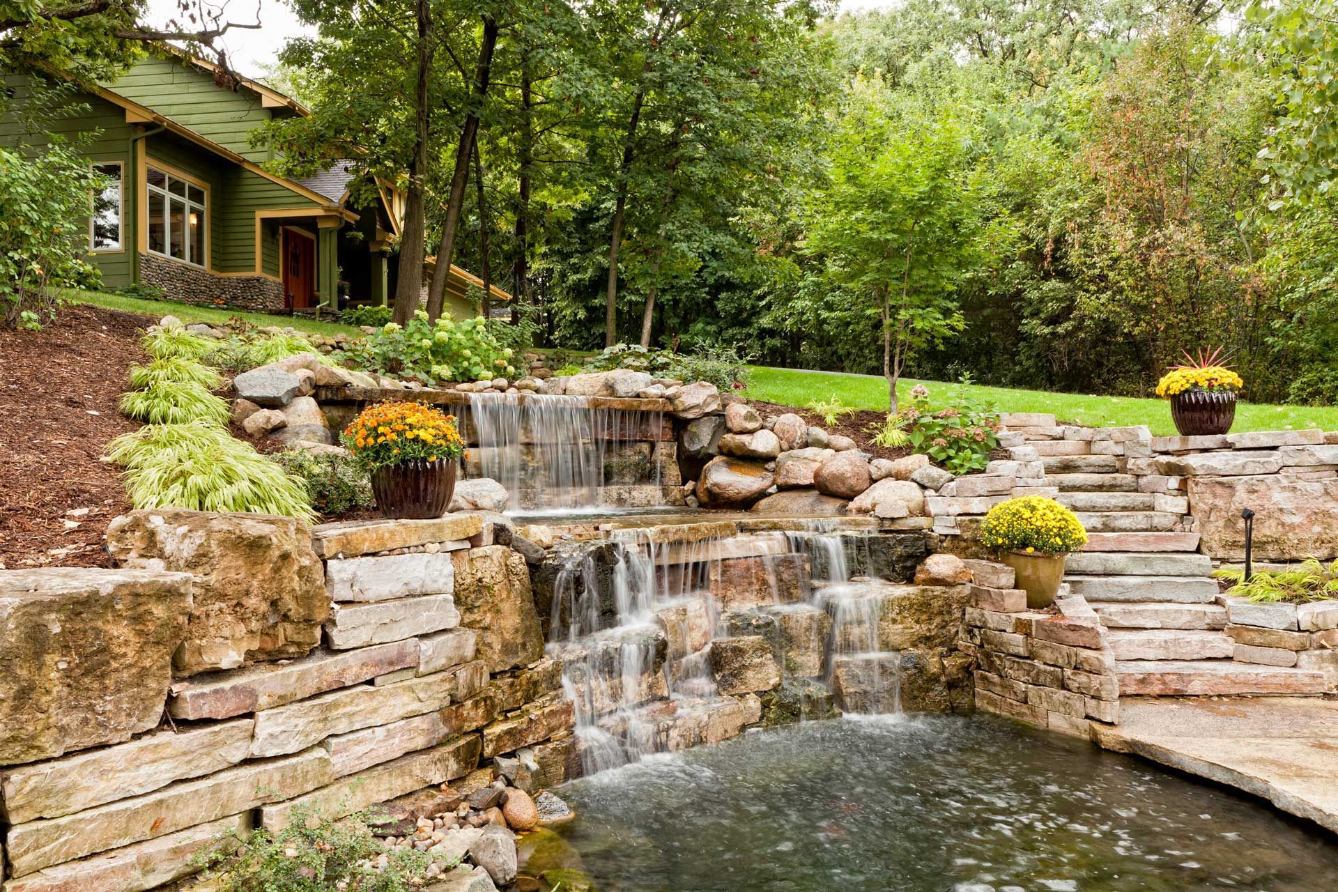 Waterfall feature with stone walls, steps, and potted plants in front of a green house.