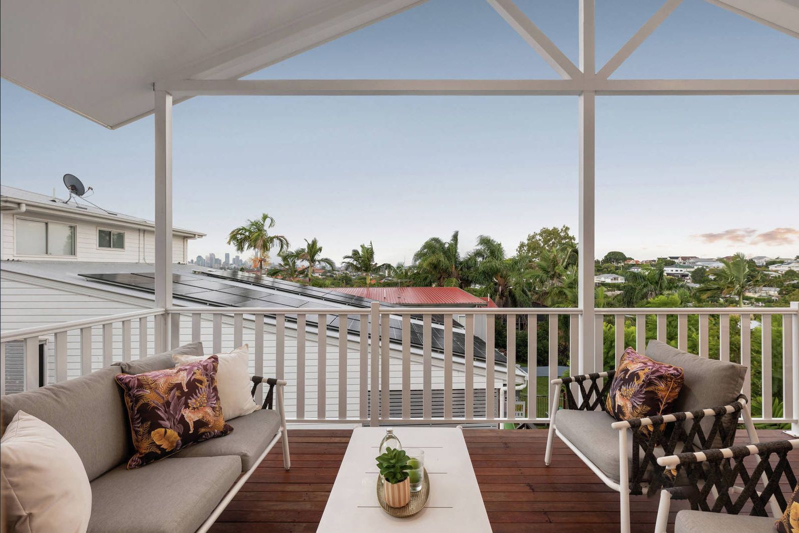 A furnished balcony with grey sofas, white railing, and a view of rooftops and trees under a clear sky.