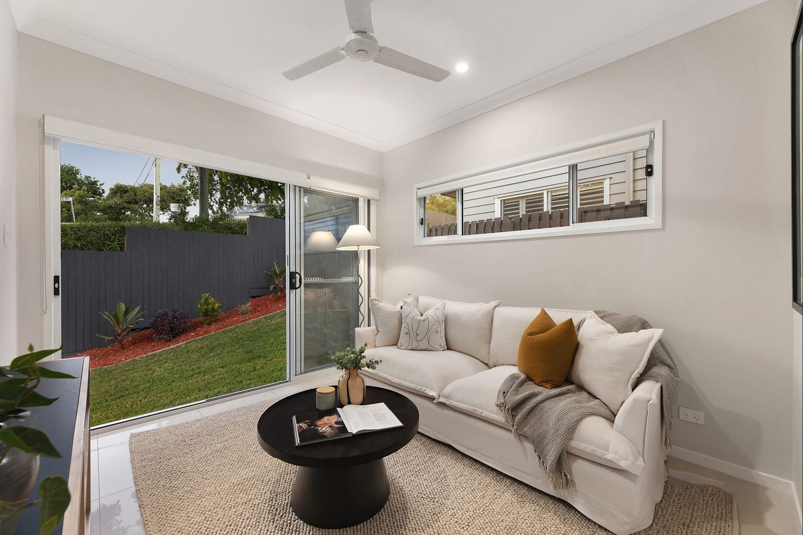 Cozy living room with white sofa, coffee table, and sliding glass door overlooking a yard with grass and a fence.