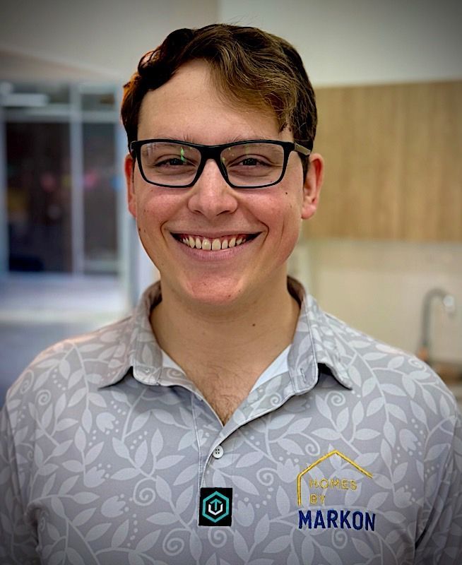 Smiling man with glasses, wearing a light gray patterned shirt. He’s indoors with a kitchen in the background. Alex Uzzell