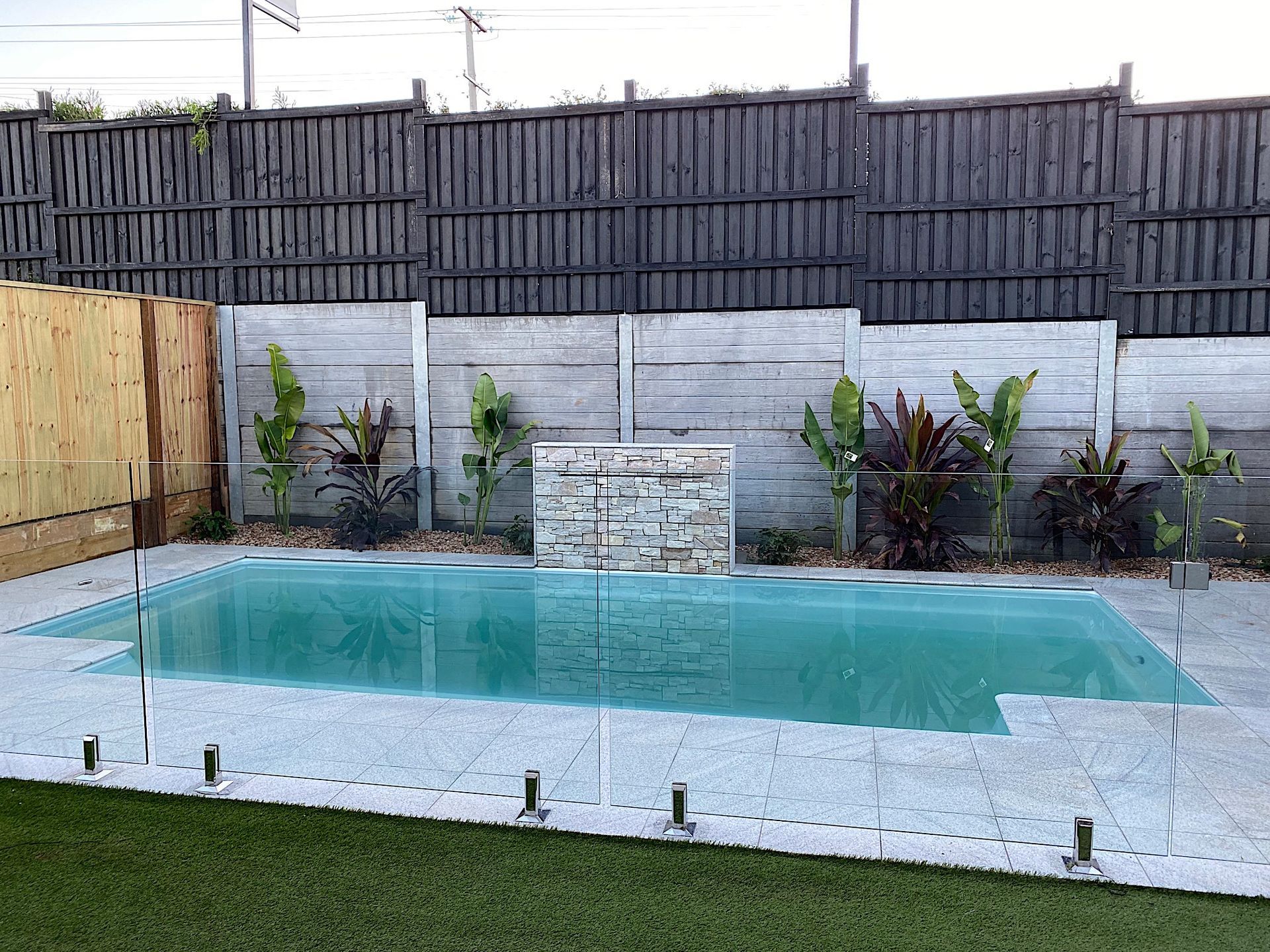 Backyard pool with glass fence, pebble surround, plants, and a stone water feature against a wooden fence.