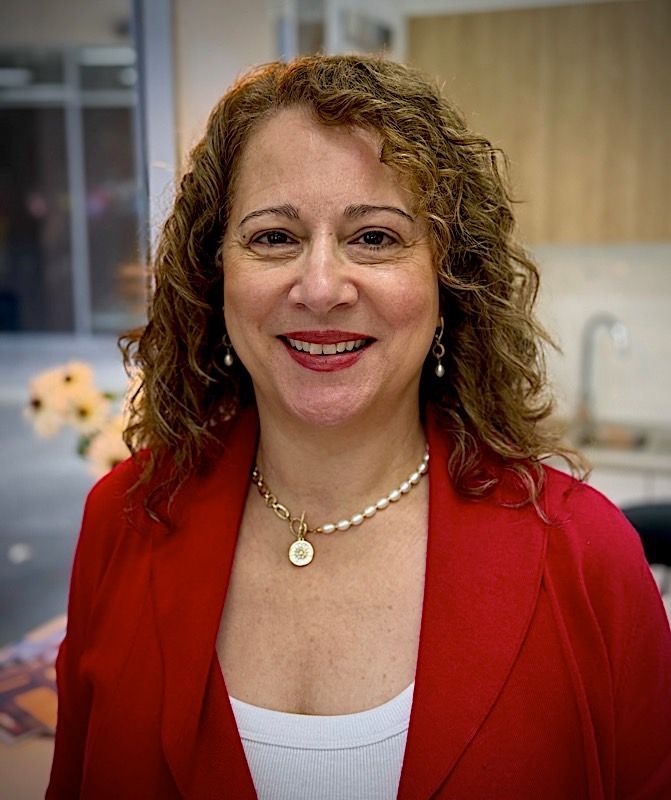 Woman with curly hair, wearing a red blazer and pearl necklace, smiles at the camera. Gianna Uzzell