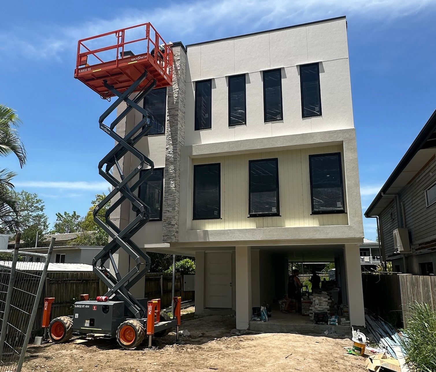 Construction of a three-story building with a scissor lift. The building is light beige with black windows. The lift is orange and gray.