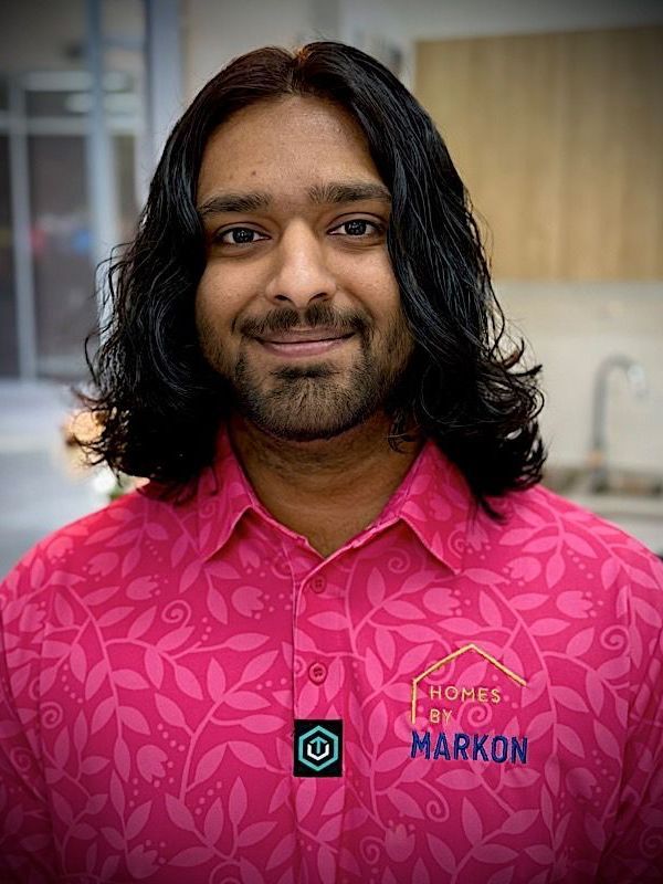 Man with long black hair, pink shirt with floral pattern, smiles in a setting, “Homes by Markon” logo visible. Jason Mathew