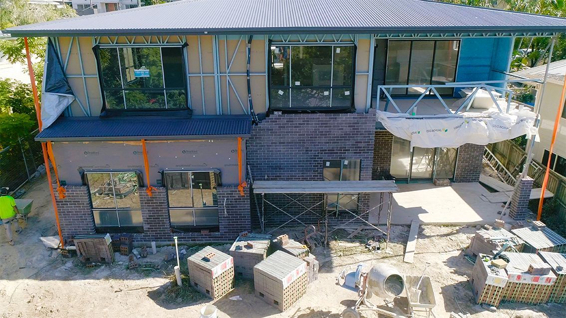 House under construction with exposed framing, brickwork, and windows, showing a partially built exterior.