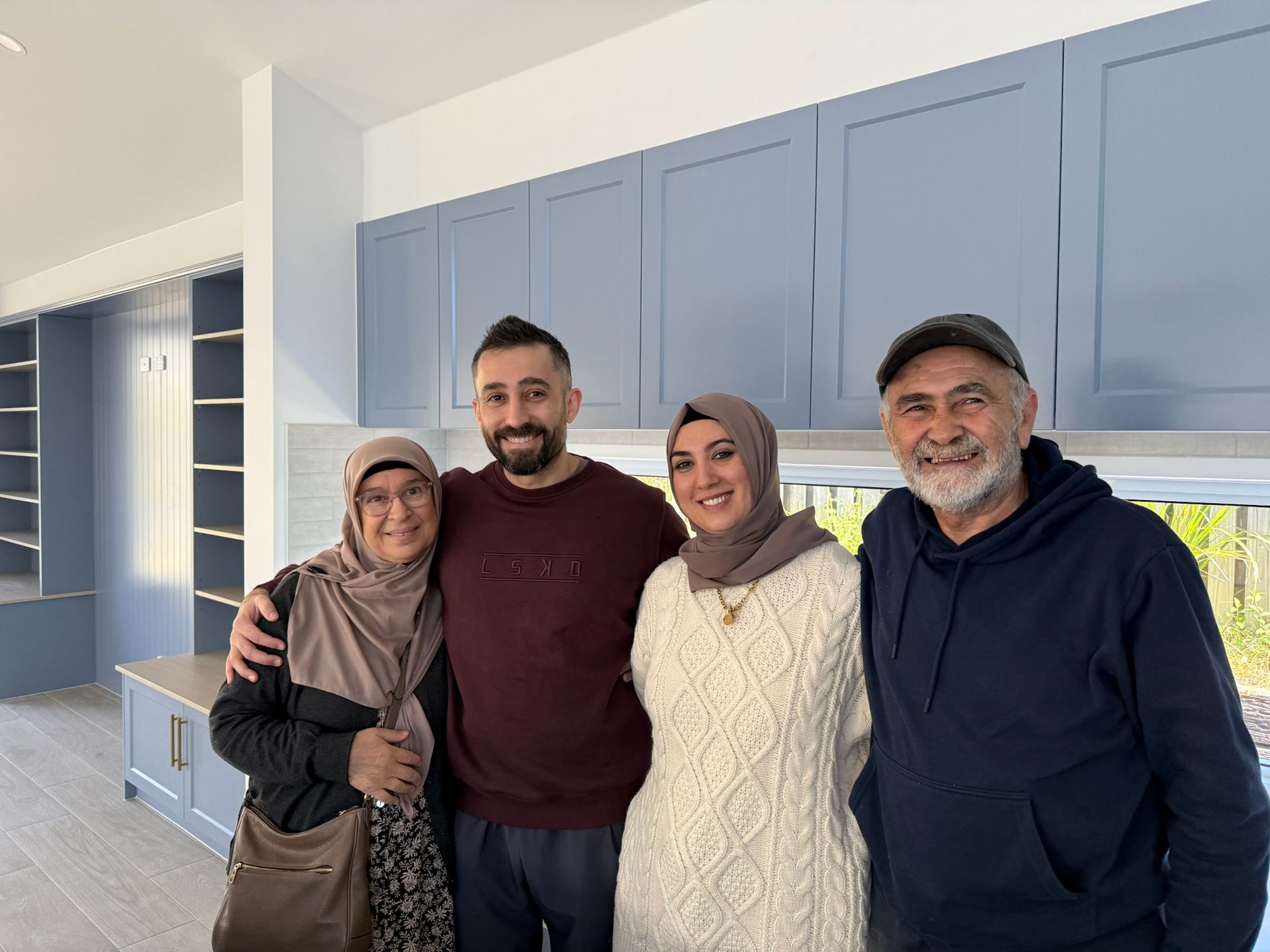 Group of six people in a kitchen, standing behind an island counter, posing. Pink and blue tones.