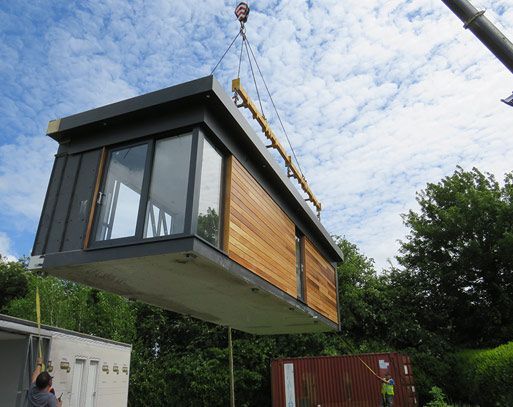 A modular building, with wooden siding and large windows, being lifted by a crane against a partly cloudy sky.