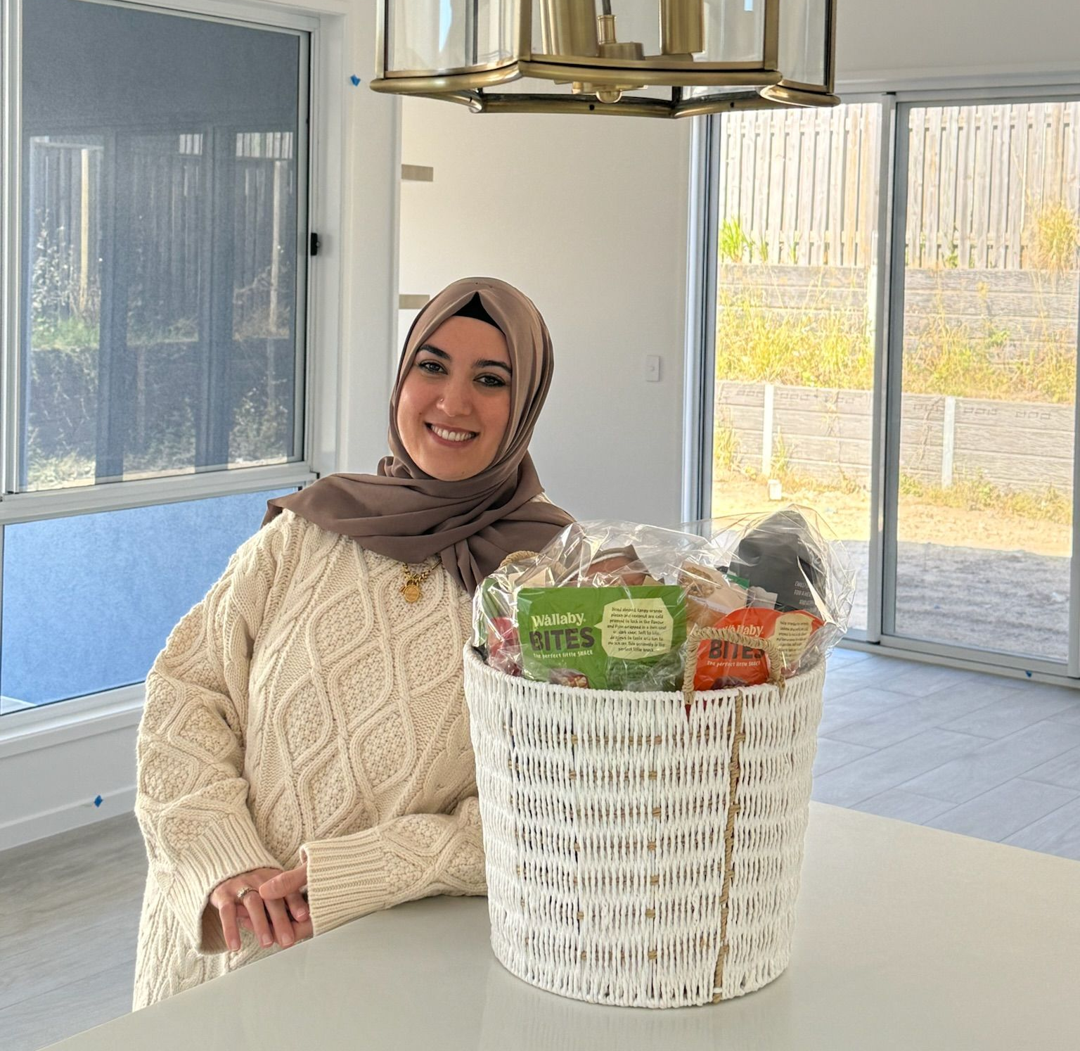 Woman in hijab smiles next to gift basket on white kitchen island, under a gold-framed light.